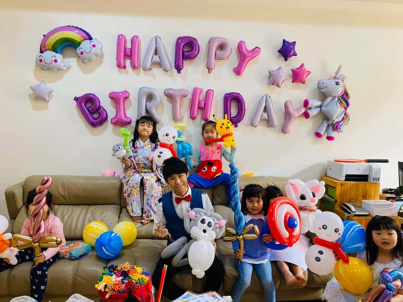 客廳牆上掛著粉紫色 HAPPY BIRTHDAY 字母氣球和獨角獸鋁箔球，柯男和孩子們坐在沙發上拿著造型氣球
