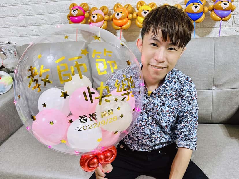 我坐在沙發上拿著透明波波球，球面金字寫著教師節快樂、香香祝賀 2022/9/28，裡面裝粉白小氣球，底部紅色蝴蝶結，後方牆上掛著一排小熊氣球