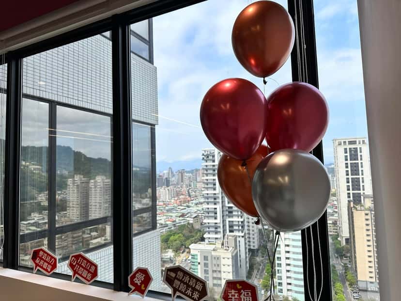 窗邊的紅色、古銅色與銀色氣球束，窗外是高樓城市景觀