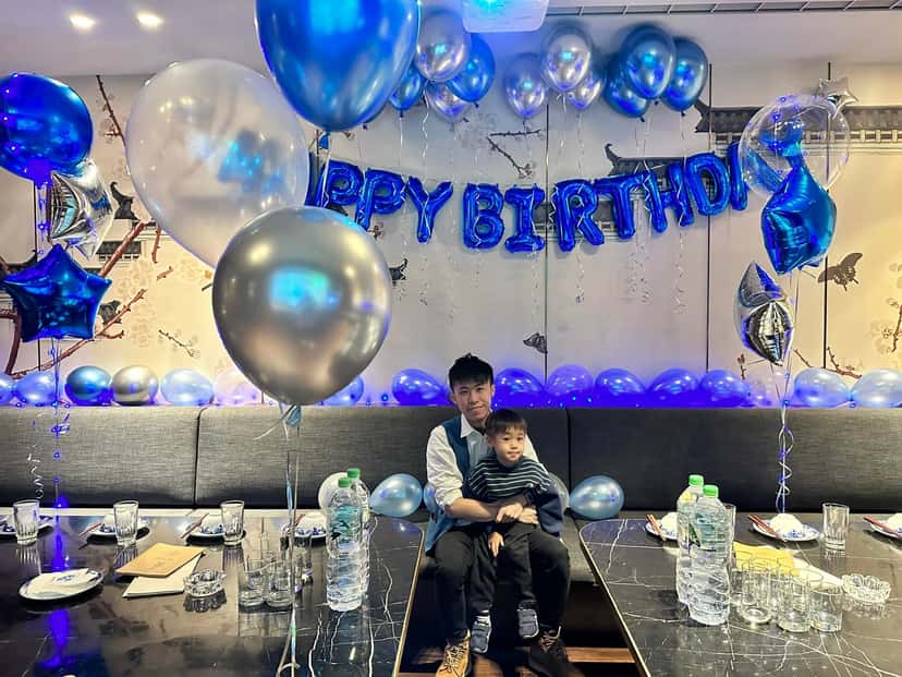 柯男抱著小男孩坐在包廂沙發上，背後是藍色 HAPPY BIRTHDAY 字母氣球、銀色和藍色氣球束