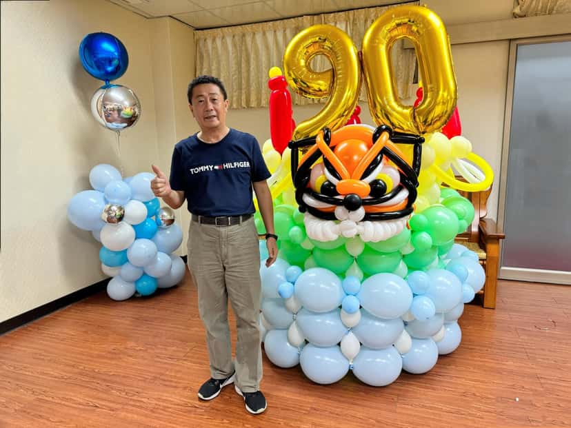 金色 90 數字氣球插在老虎頭氣球塔上，底下是藍白氣球，一位長輩站在旁邊比讚