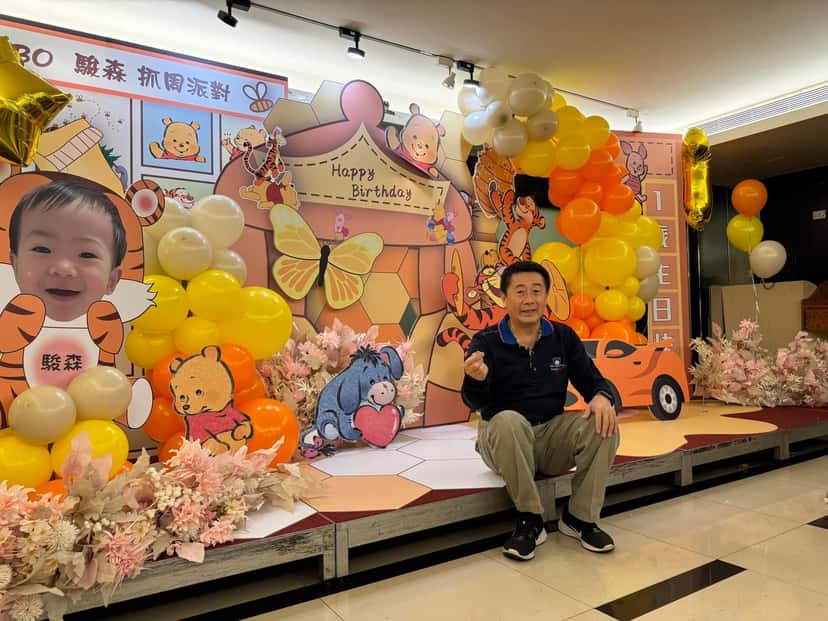 小熊維尼抓周舞台全景，左邊是寶寶大頭立牌，中間 Happy Birthday 背板，兩側黃橘氣球串和跑車立牌，長輩坐在台上