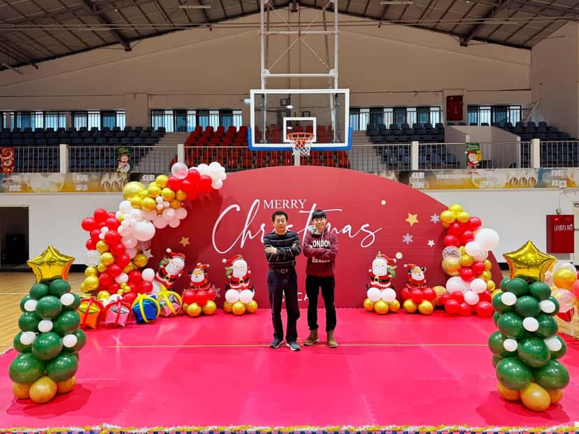 體育館舞台上的紅色 Merry Christmas 背板，兩側是紅白金氣球、聖誕老公公和綠金聖誕樹氣球柱，兩人站在中間