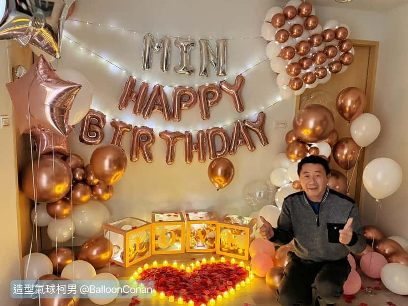 玫瑰金 HAPPY BIRTHDAY 字母氣球牆下，地上是玫瑰花瓣與蠟燭排成的愛心，長輩蹲在旁邊比讚