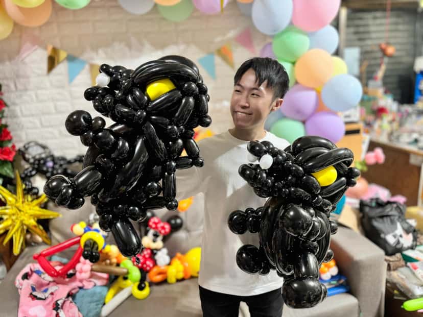 柯男在工作室裡雙手各拿一隻全黑黃眼的哥吉拉造型氣球