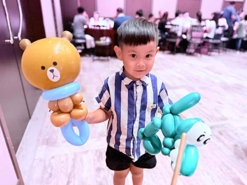 穿條紋襯衫的小男孩一手拿咖啡色小熊、一手拿綠色小狗造型氣球