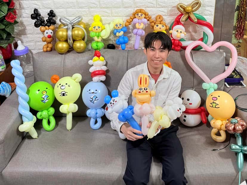 柯男坐在沙發上抱著一把造型氣球，周圍擺滿雪人、公主娃娃、愛心和各種角色氣球