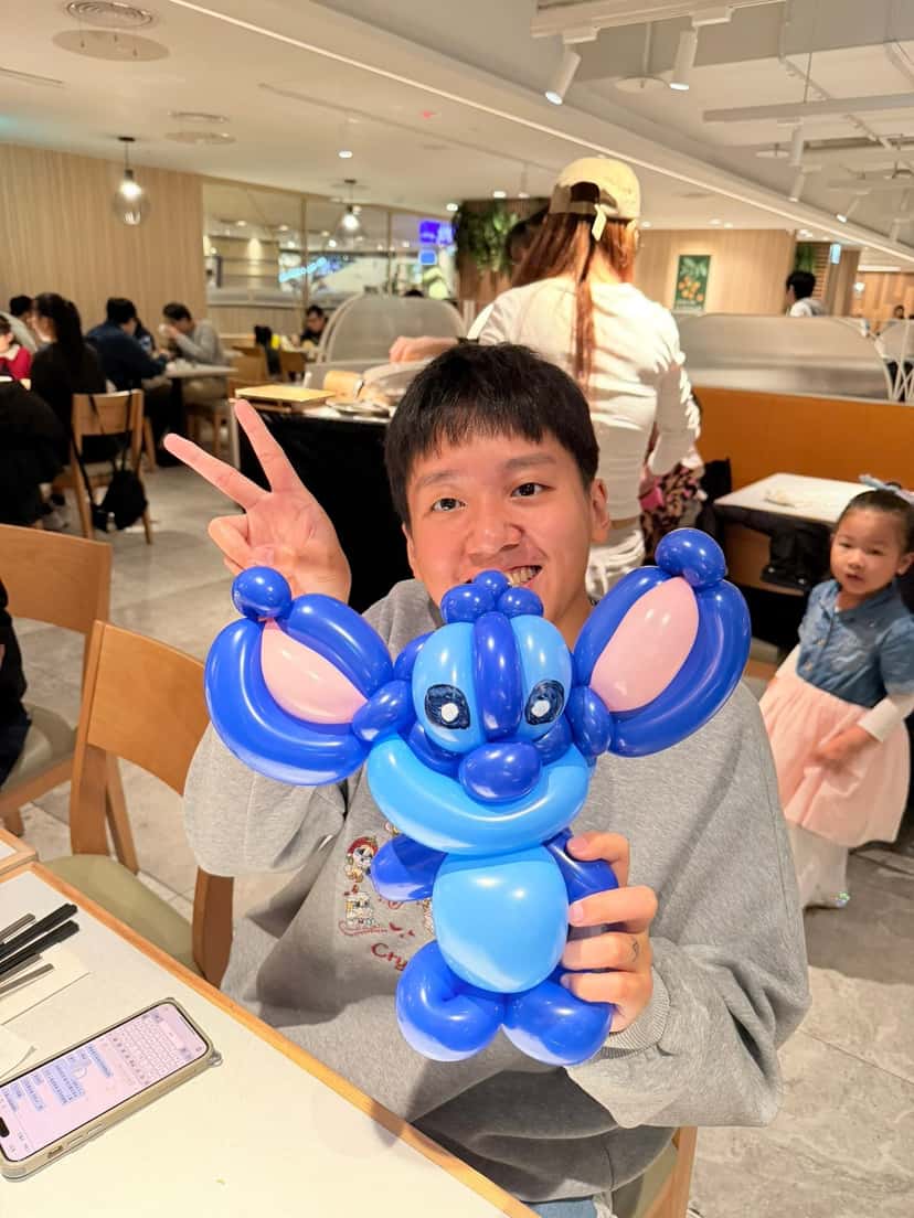 大人坐在餐桌旁比 YA，雙手捧著藍色史迪奇造型氣球