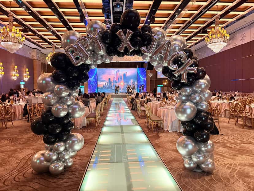 宴會廳伸展台入口的黑銀氣球拱門，拱門上方排著銀色鋁箔字母氣球，遠處是大型螢幕舞台