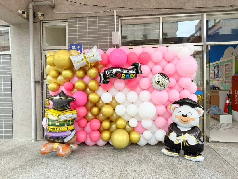 校舍門口的粉白金色氣球牆，掛著 Congratulations GRAD 鋁箔字條，旁邊有穿畢業袍的熊與書本造型氣球