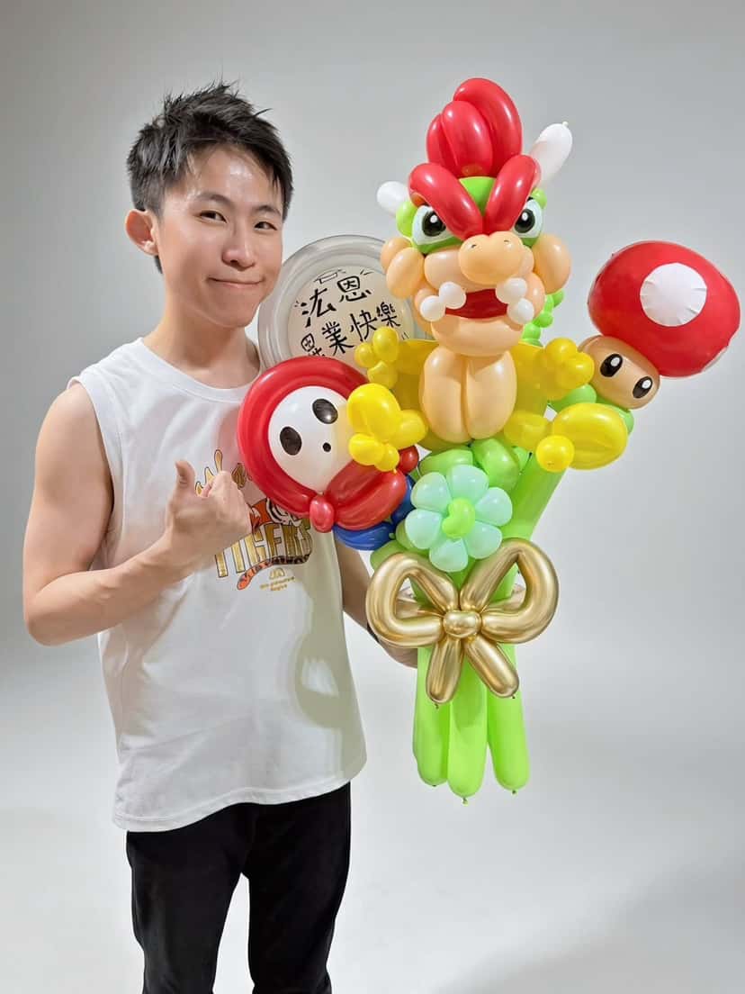 穿白色背心的柯男比讚，手抱庫巴氣球花束，花束後方圓框貼著畢業快樂客製文字
