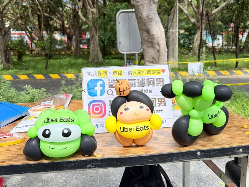 新北市監理所戶外攤位桌上擺著三款 Uber Eats 造型氣球：綠色大眼小車、黃色圍兜娃娃與綠黑機車，都貼有品牌貼紙