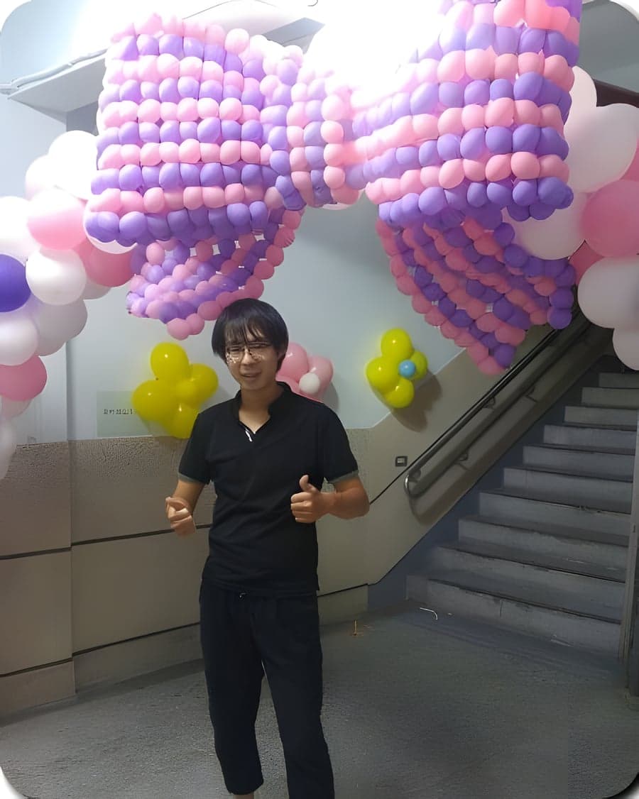 一位穿黑衣的男士站在編織蝴蝶結氣球拱門下比讚