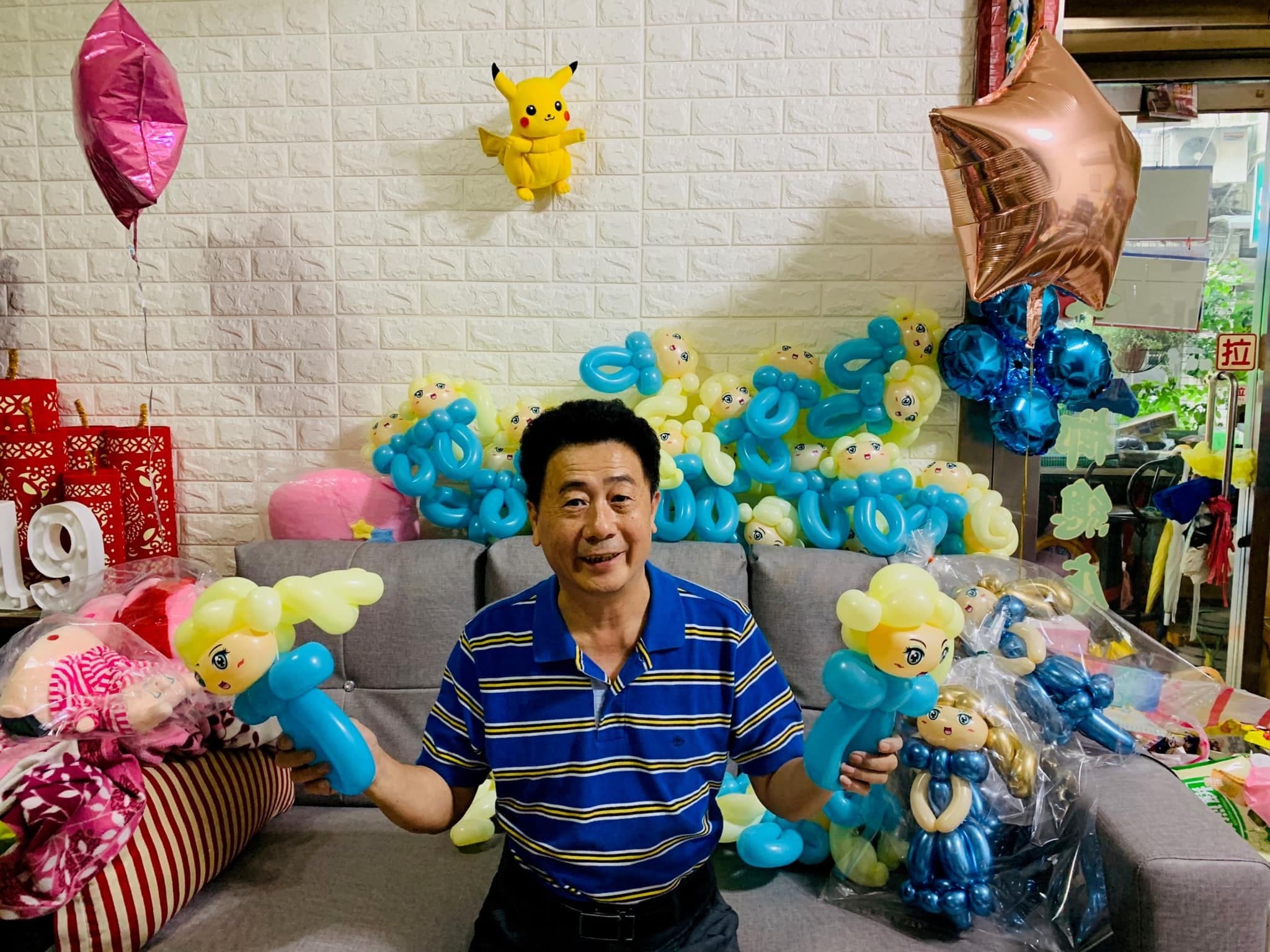 柯爸坐在沙發前，兩手拿著艾莎造型氣球，後方堆滿成品