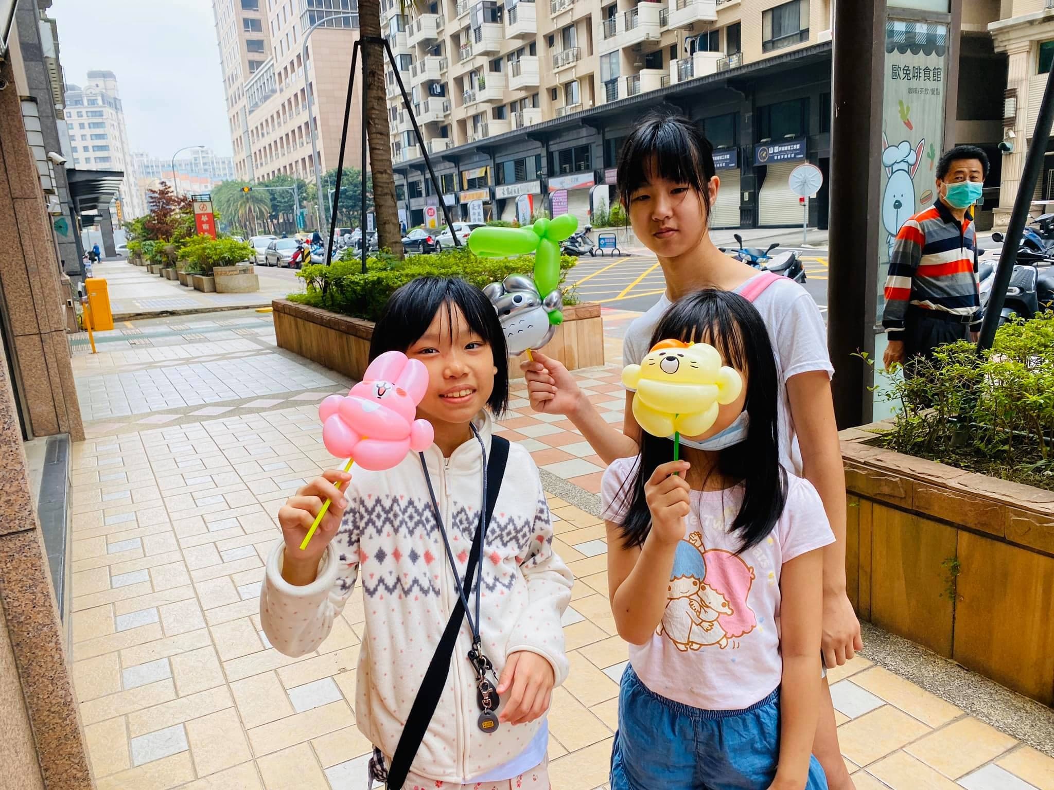 三個女孩在人行道上舉著卡娜赫拉粉紅兔兔、黃色獅子與綠色角色氣球