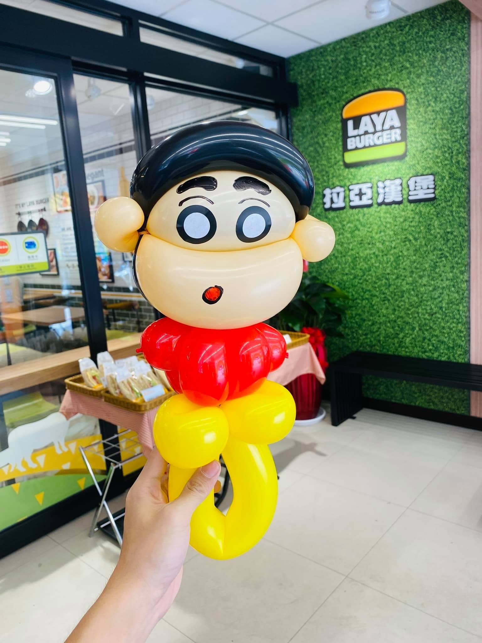 手上拿著蠟筆小新造型氣球，背景是拉亞漢堡店內的綠色招牌牆