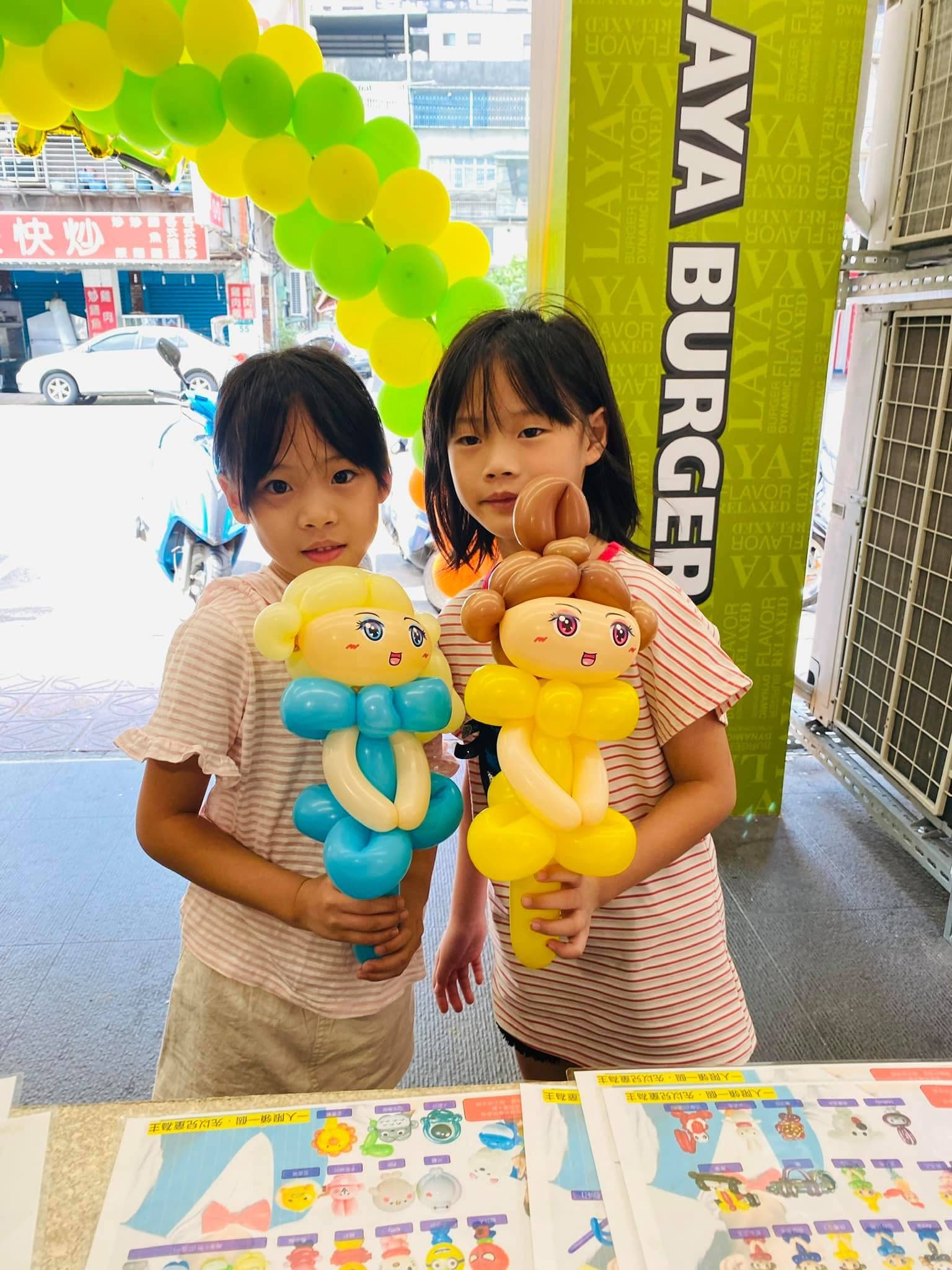 兩個女孩在拉亞漢堡門口，各拿一隻淺藍衣和黃衣的公主娃娃造型氣球