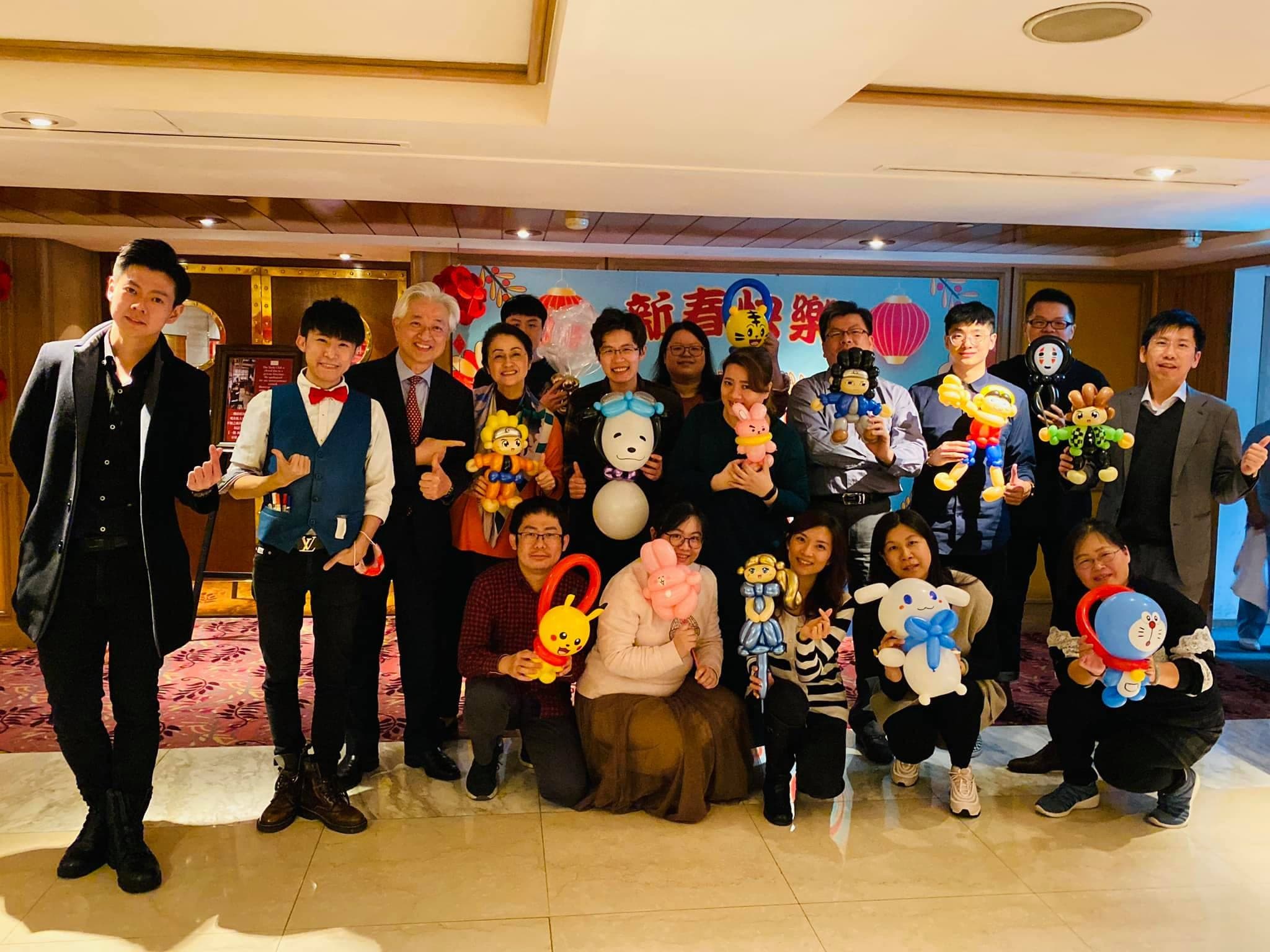 春酒會場大合照，大人們拿著各式造型氣球站在新春布條前