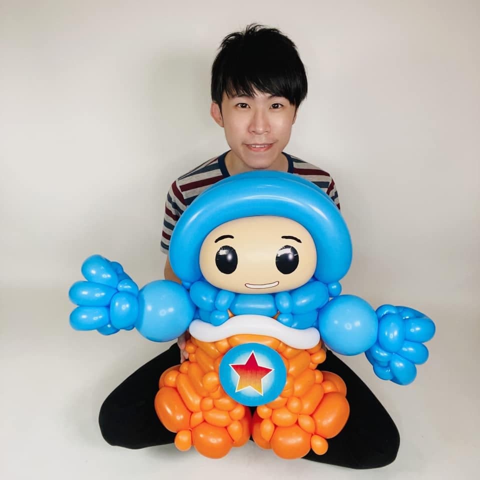 柯男坐在白色背景前，懷裡是全球探險隊凱恩造型氣球