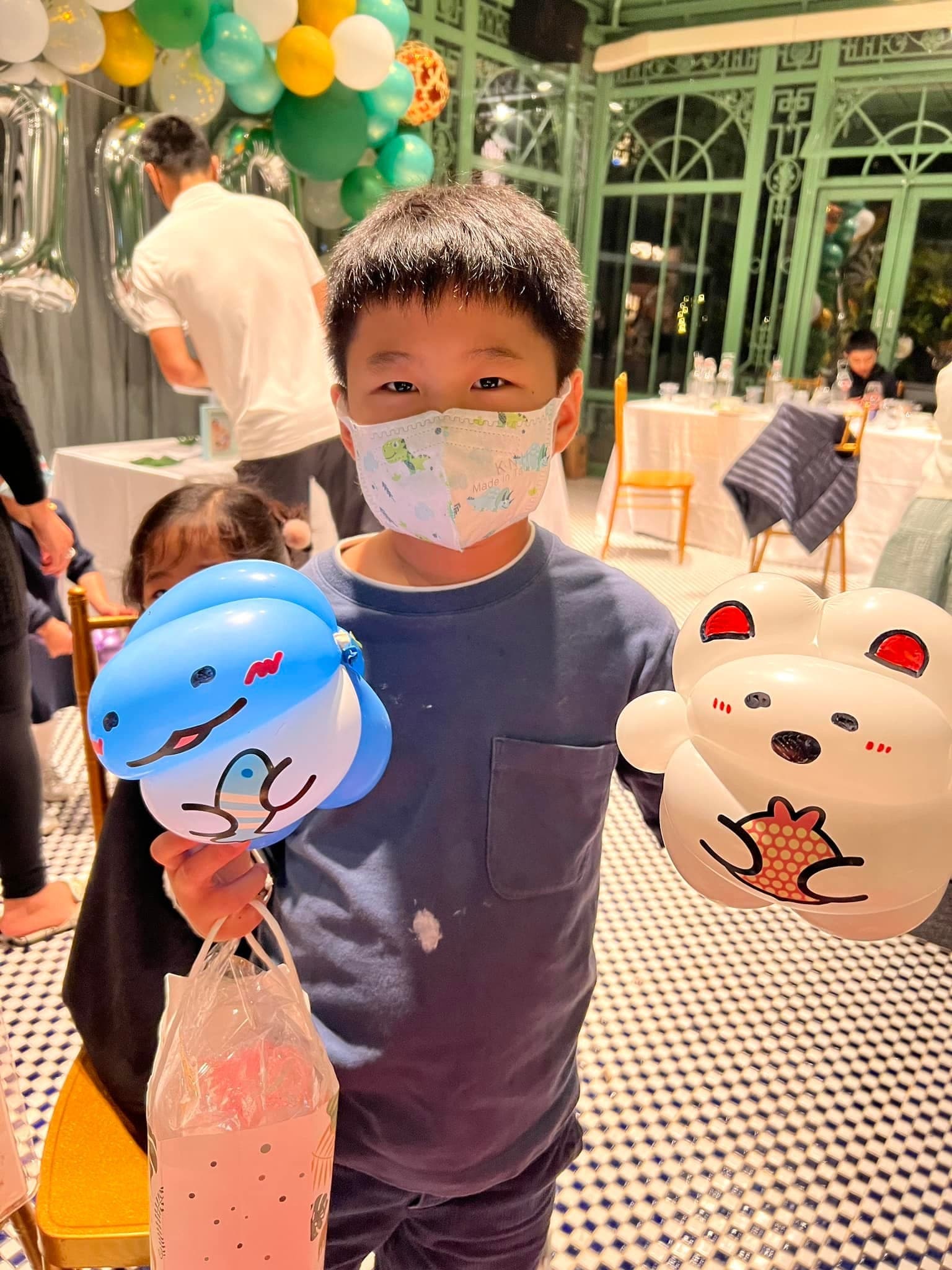 小男孩一手拿藍色小鯊魚氣球、一手拿白色小熊氣球，後方是玻璃溫室風格的餐廳