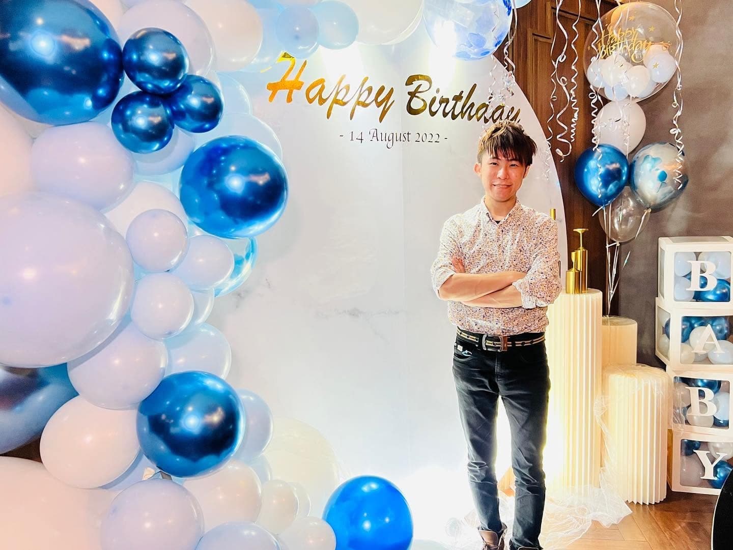 柯男雙手交叉站在藍白氣球拱與 Happy Birthday 圓形背板前