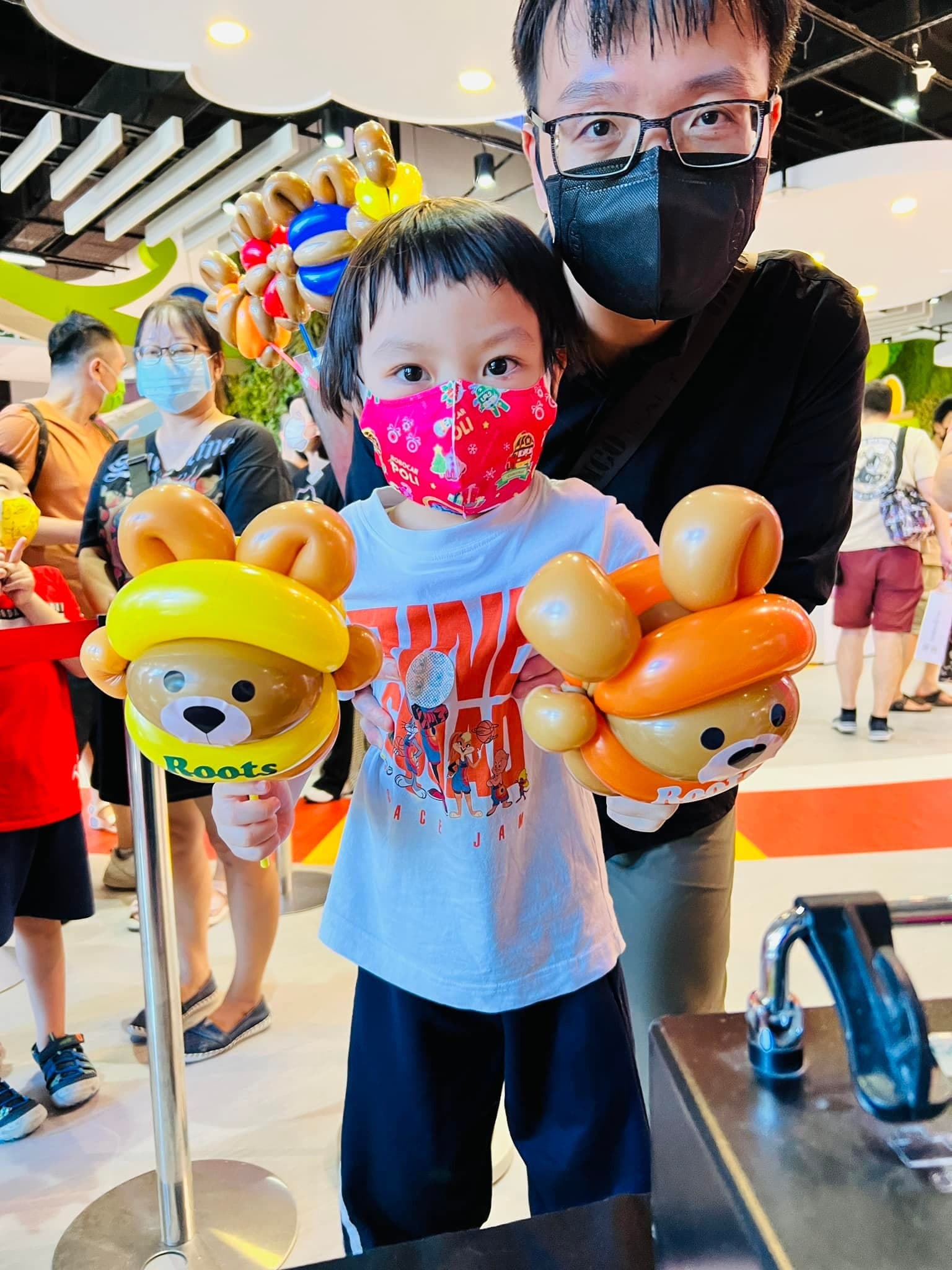 戴紅色口罩的孩子拿著黃帽和橘帽海狸寶寶氣球，爸爸在後面陪著