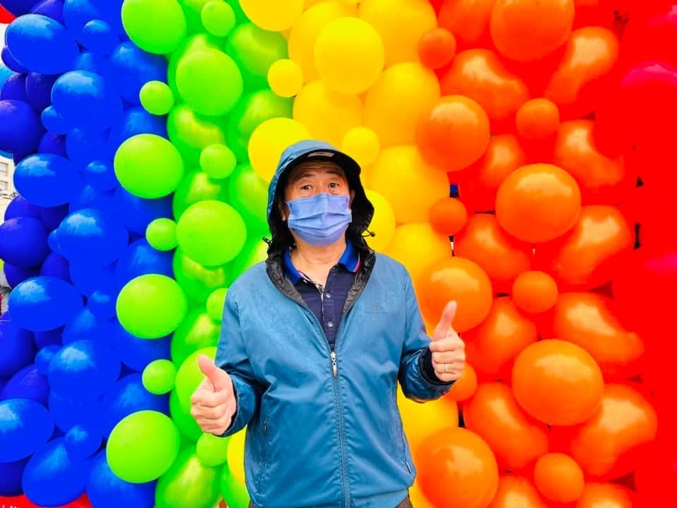 穿藍色外套的男士在藍、綠、黃、橘、紅色漸層彩虹氣球牆前比讚
