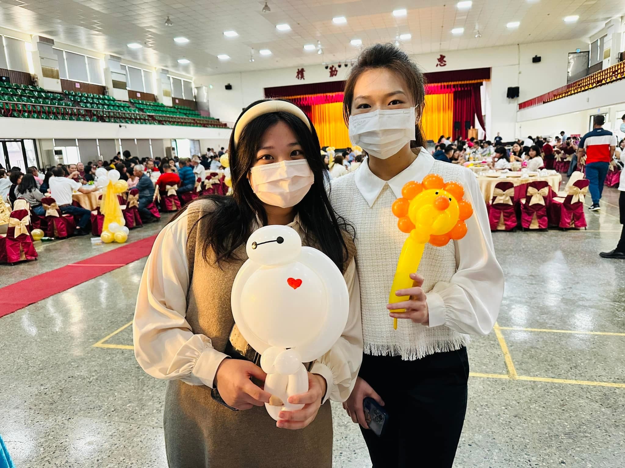 兩位女賓客在婚宴會場合照，一人抱白色杯麵氣球、一人拿橘黃色獅子氣球棒