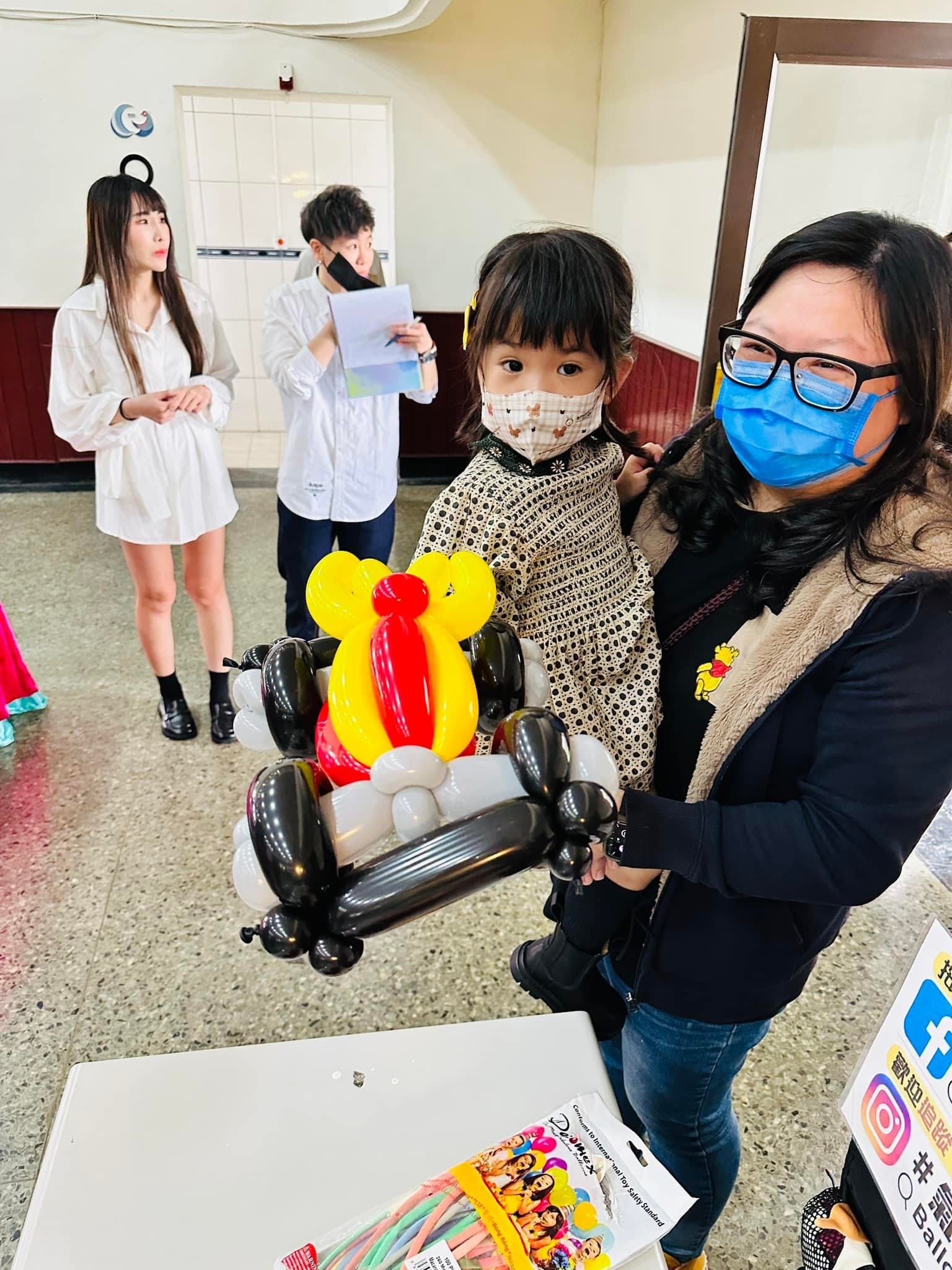 媽媽抱著小女孩，手上拿著黑色輪子、黃紅色車身的機車造型氣球