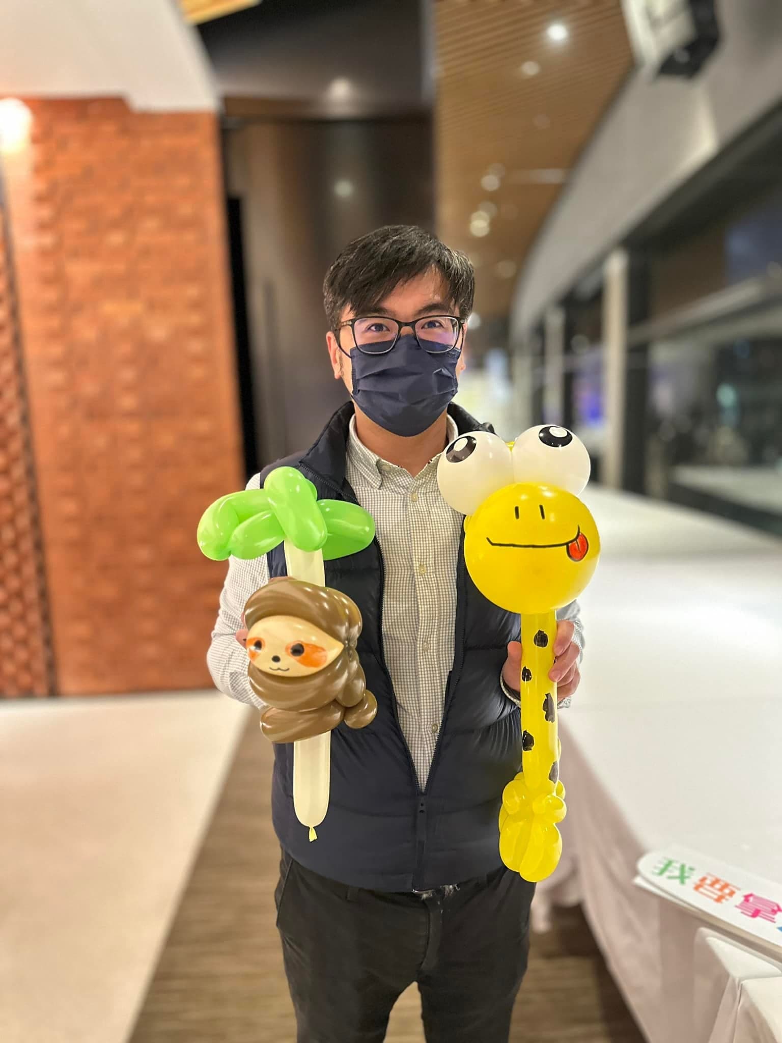 戴眼鏡的男賓客一手拿樹懶氣球、一手拿吐舌頭的長頸鹿氣球