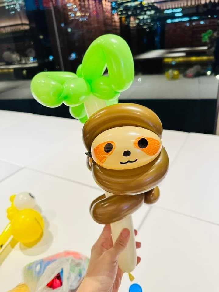 咖啡色樹懶造型氣球抱著綠葉樹幹，橘色眼圈