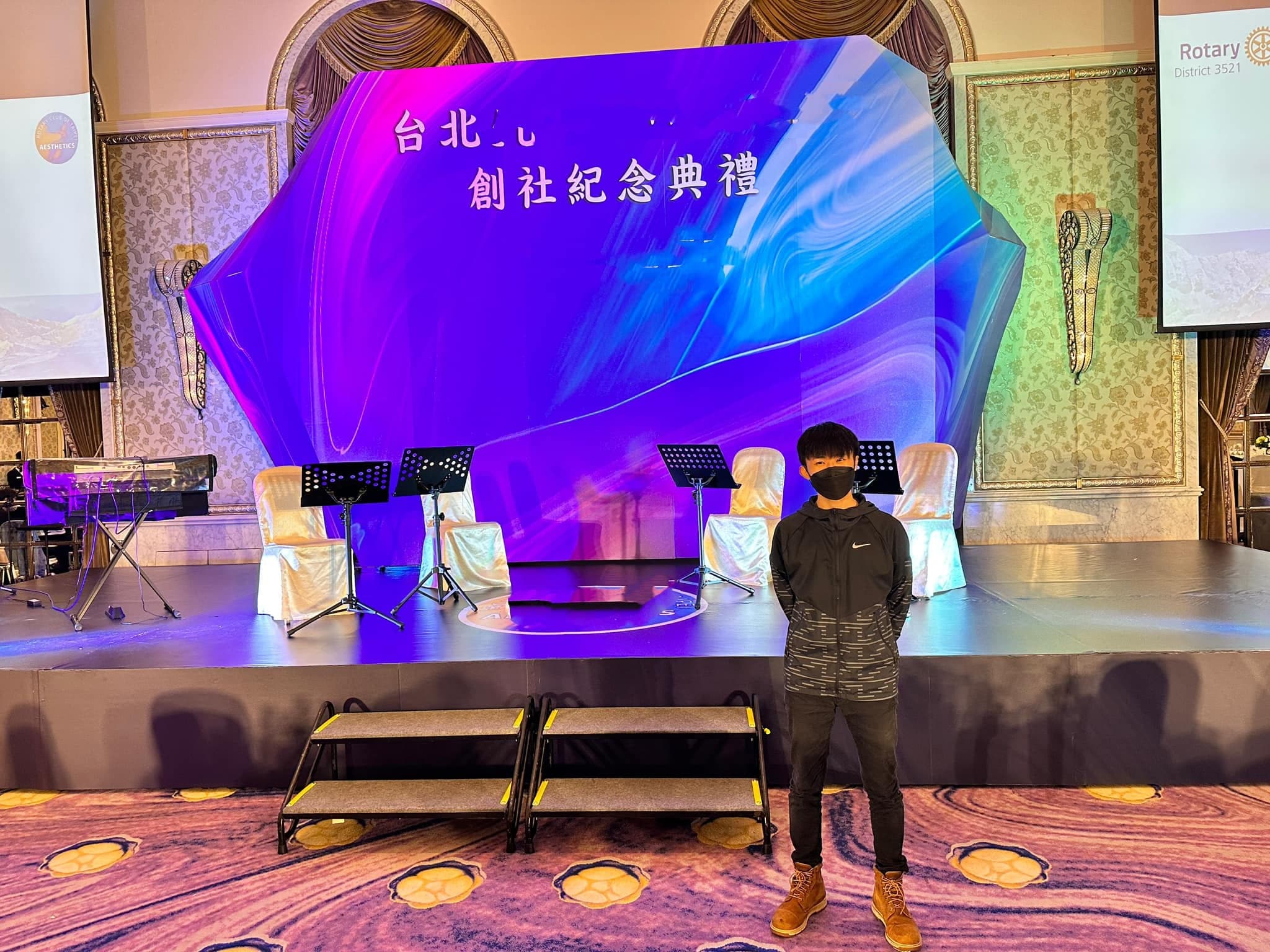 柯男站在立體鑽石背板舞台前，舞台上擺著譜架與白色椅子
