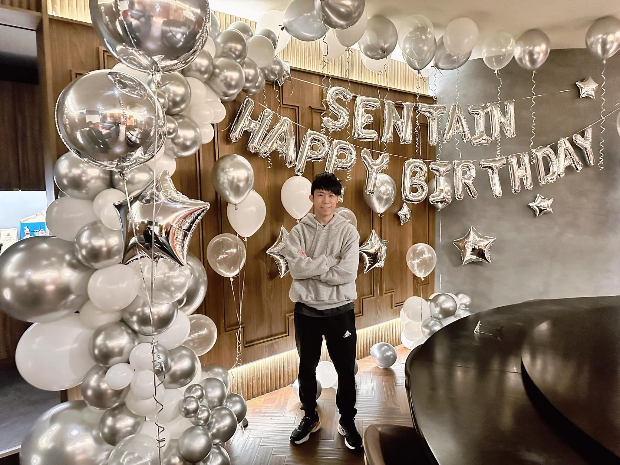 柯男雙手抱胸站在銀白色氣球串與 HAPPY BIRTHDAY 字母氣球前