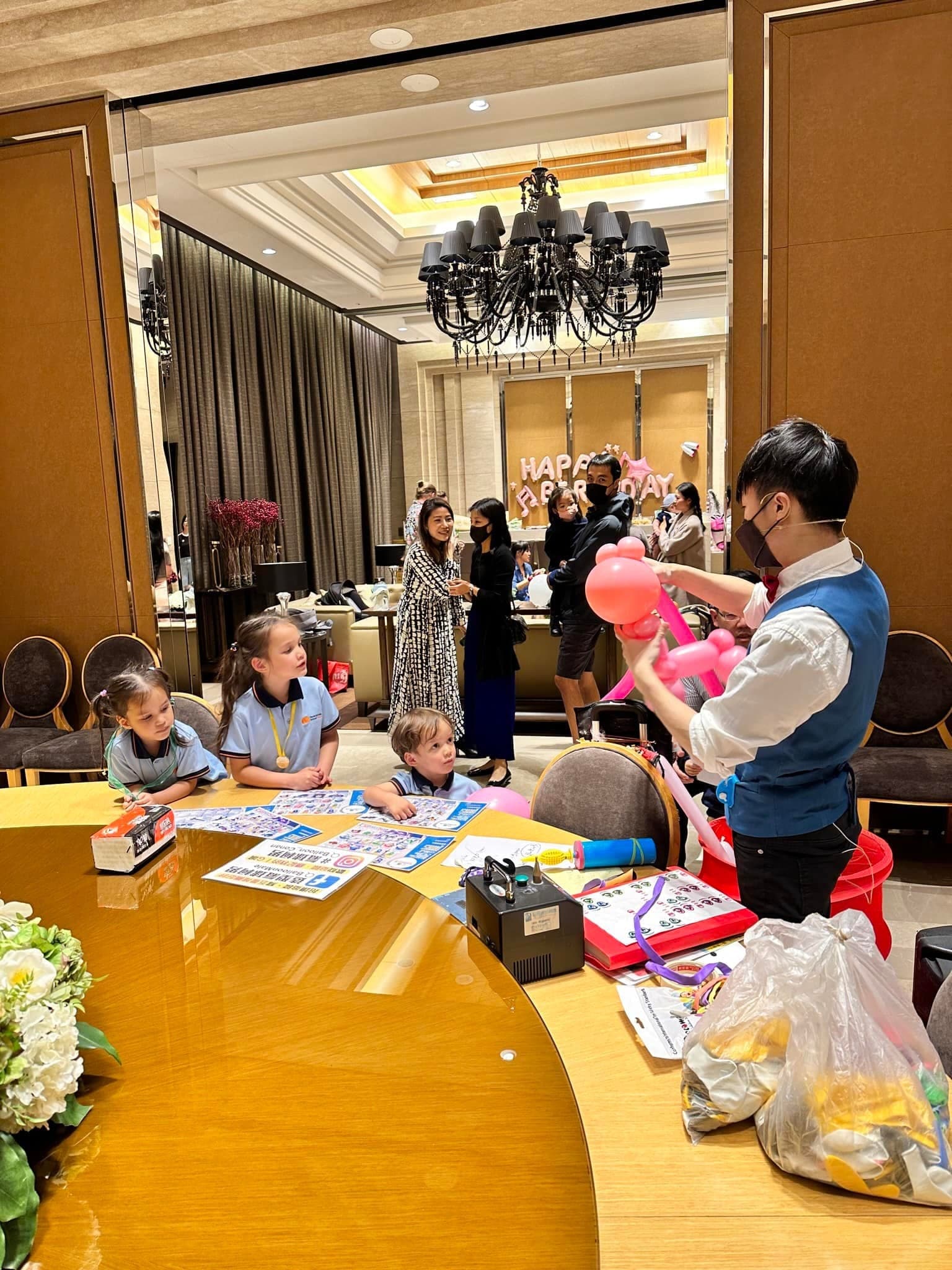 水晶吊燈下的派對廳，柯男在圓桌旁替孩子折粉紅色氣球