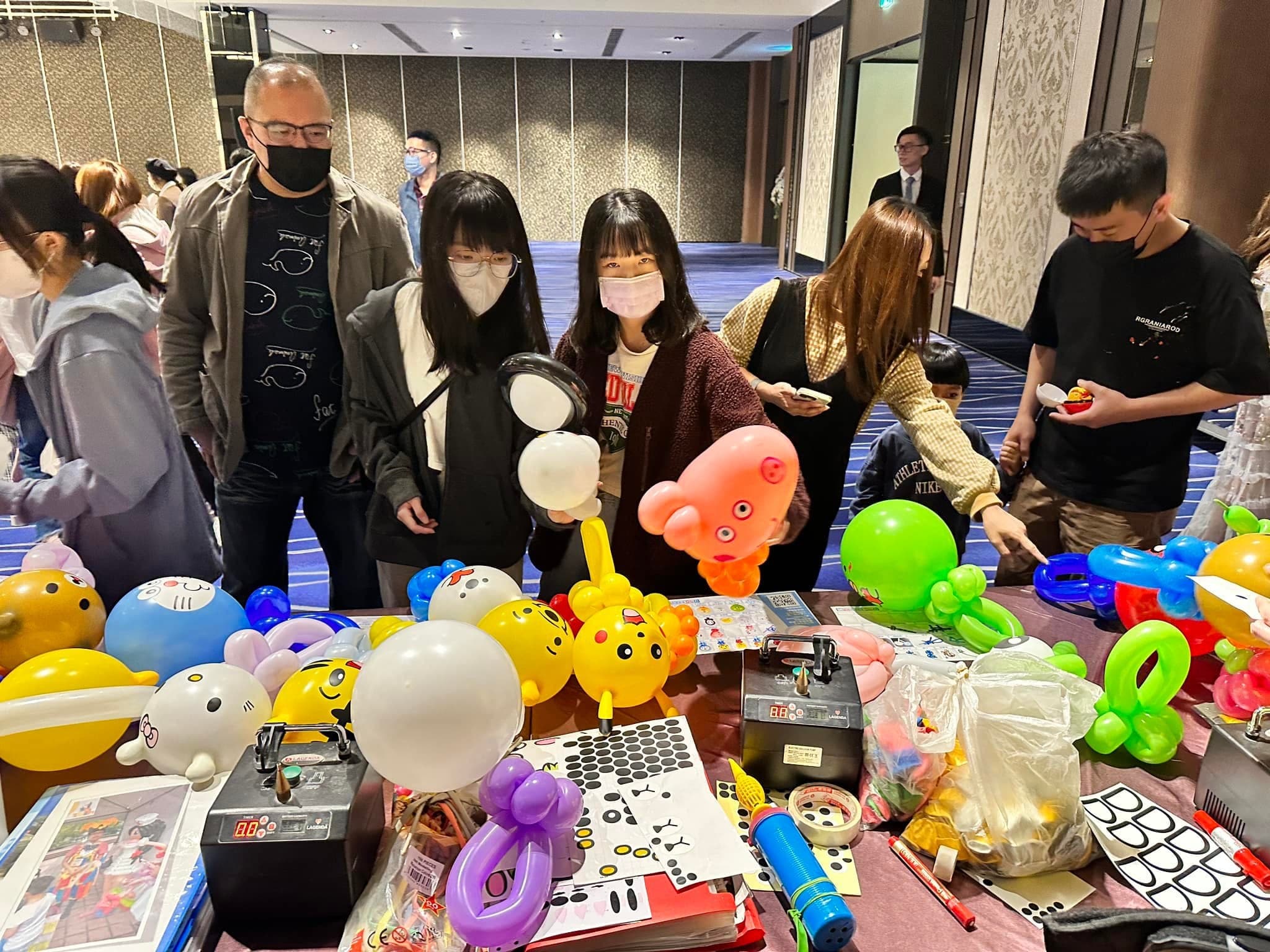 春酒賓客圍在折氣球桌前挑選造型，桌上有哆啦A夢與粉紅章魚氣球