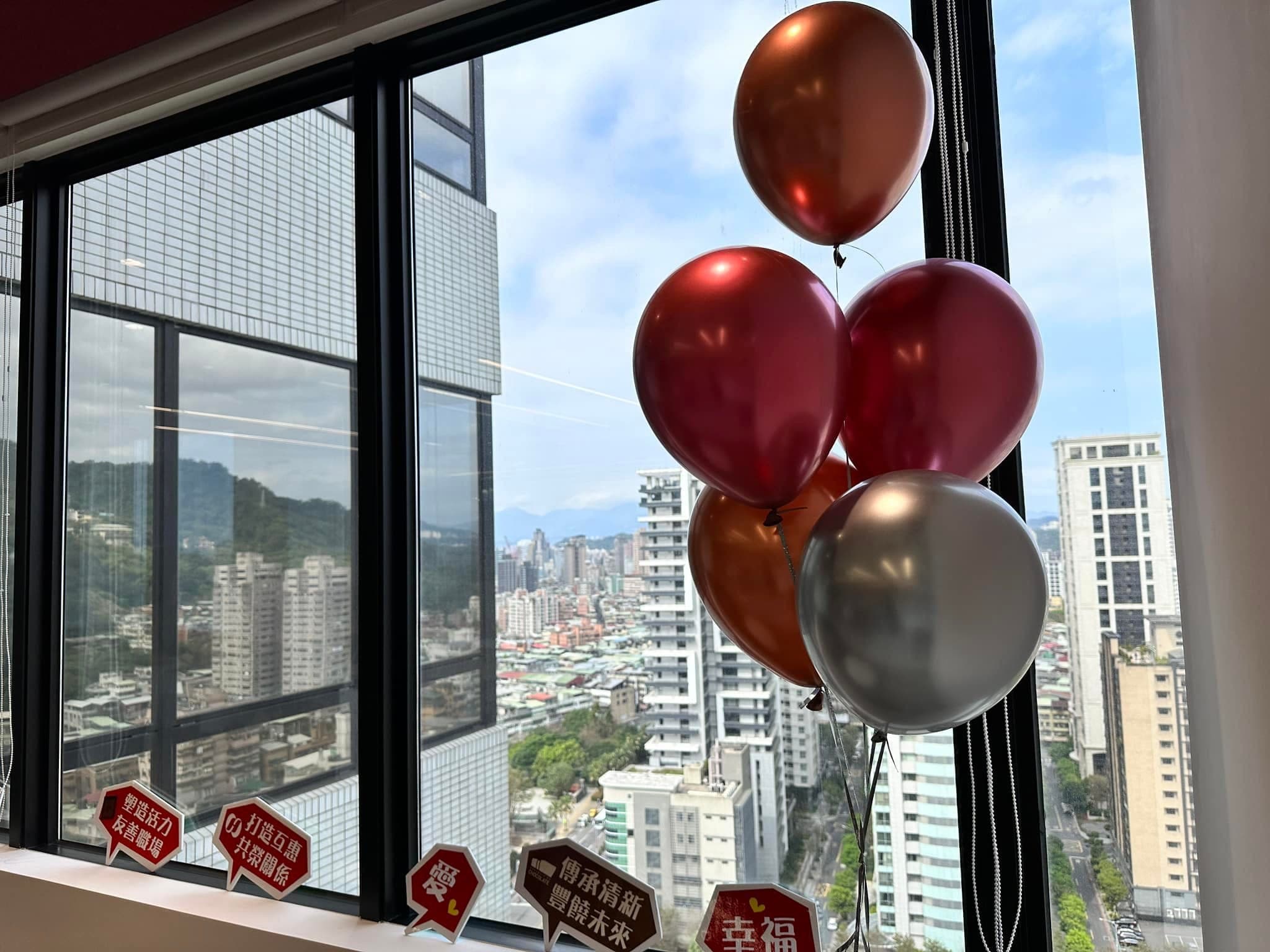 窗邊的紅色、古銅色與銀色氣球束，窗外是高樓城市景觀
