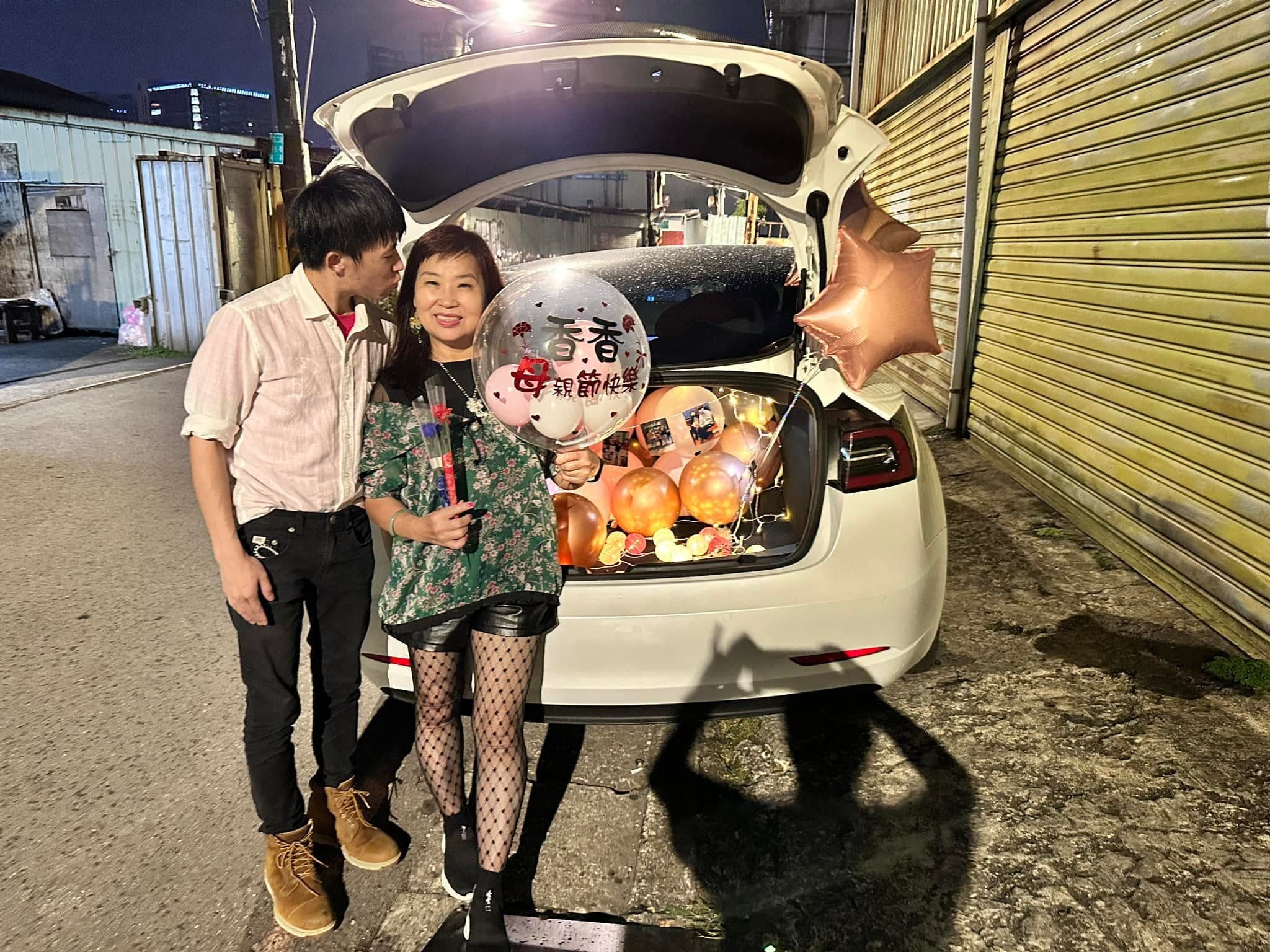 柯男和媽媽站在打開的後車廂旁，媽媽拿著寫字的透明波波球，車廂裡是玫瑰金與香檳色氣球