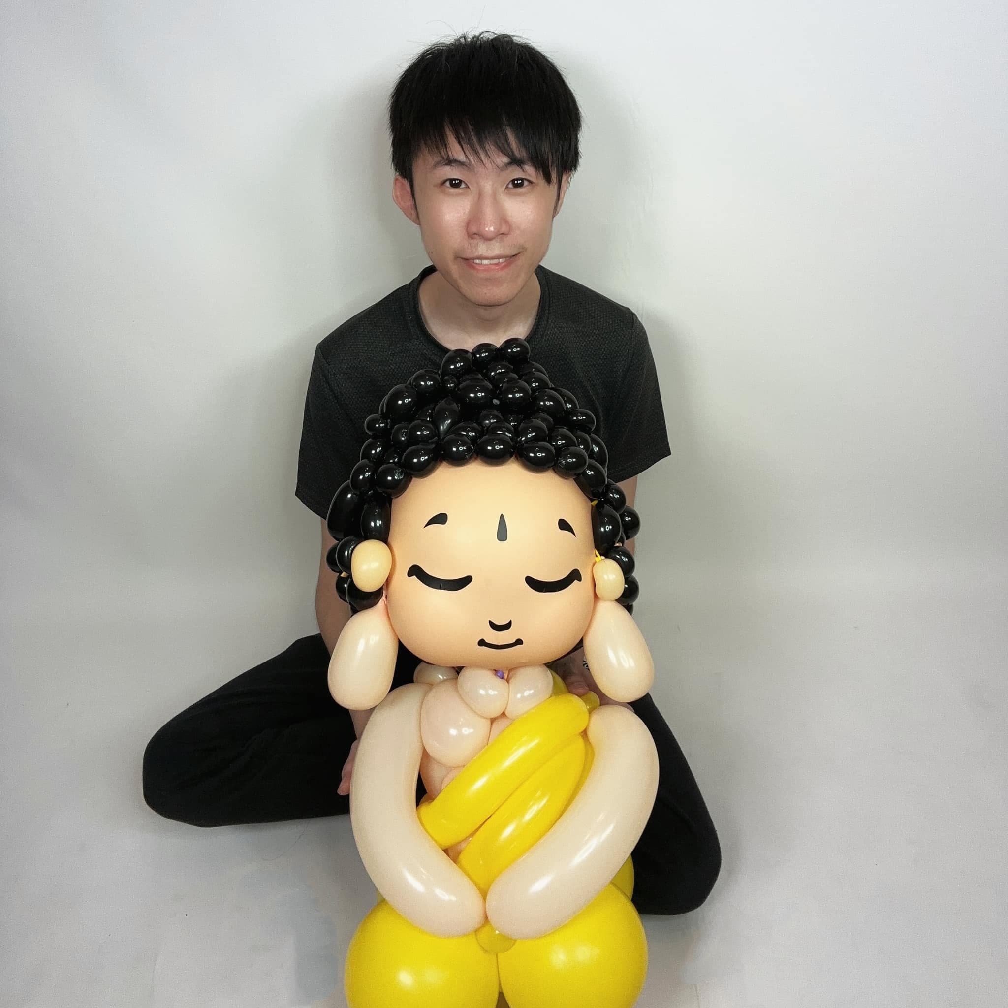 柯男坐在白色背景前，抱著完成的釋迦牟尼佛氣球人偶