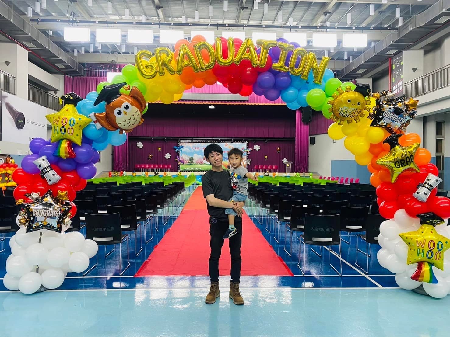 柯男抱著小男孩站在禮堂紅地毯上，頭頂是 GRADUATION 字母彩虹拱門與貓頭鷹、星星氣球