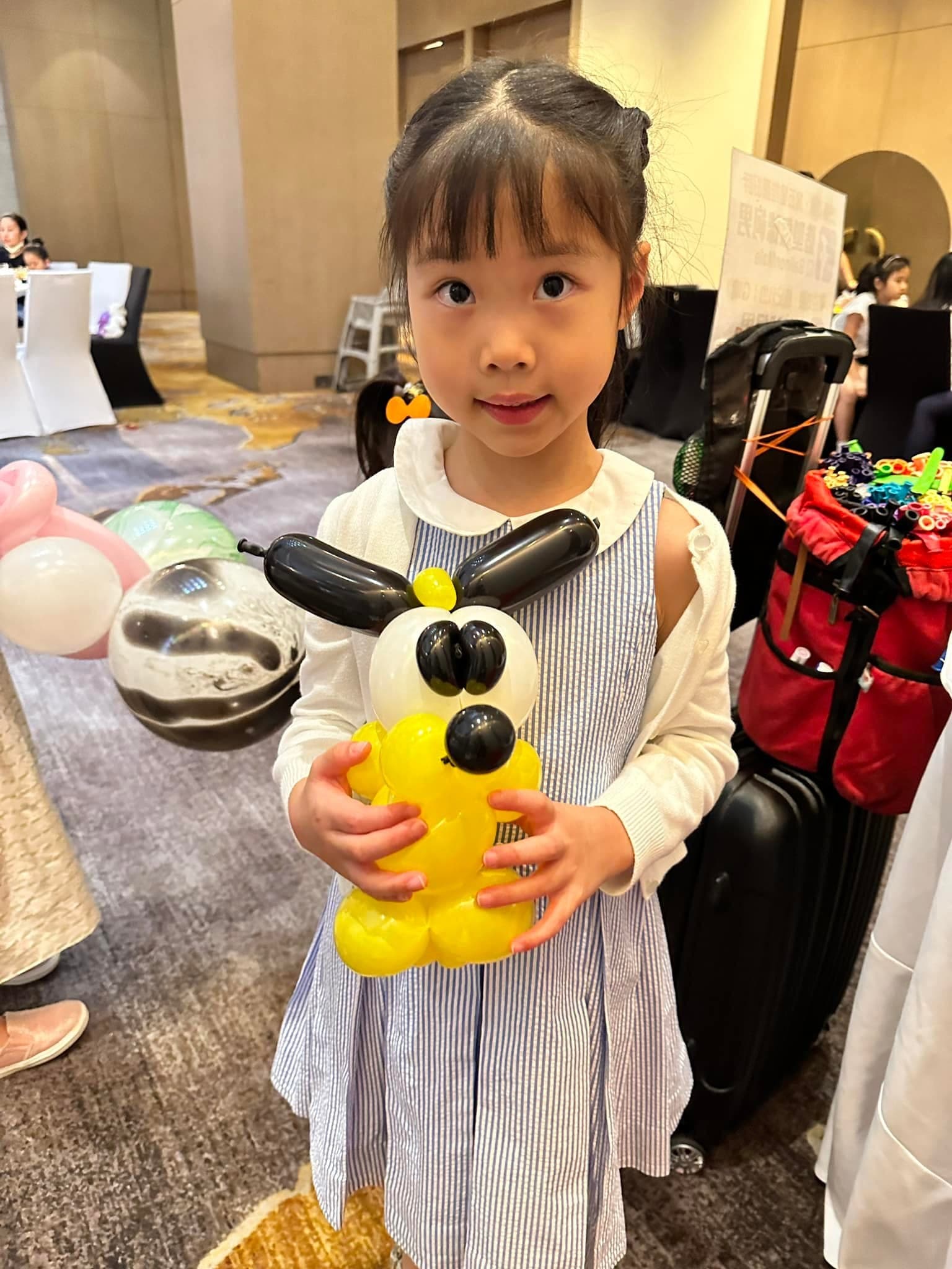 小女孩抱著黑耳朵的黃色小狗造型氣球，站在宴會廳裡