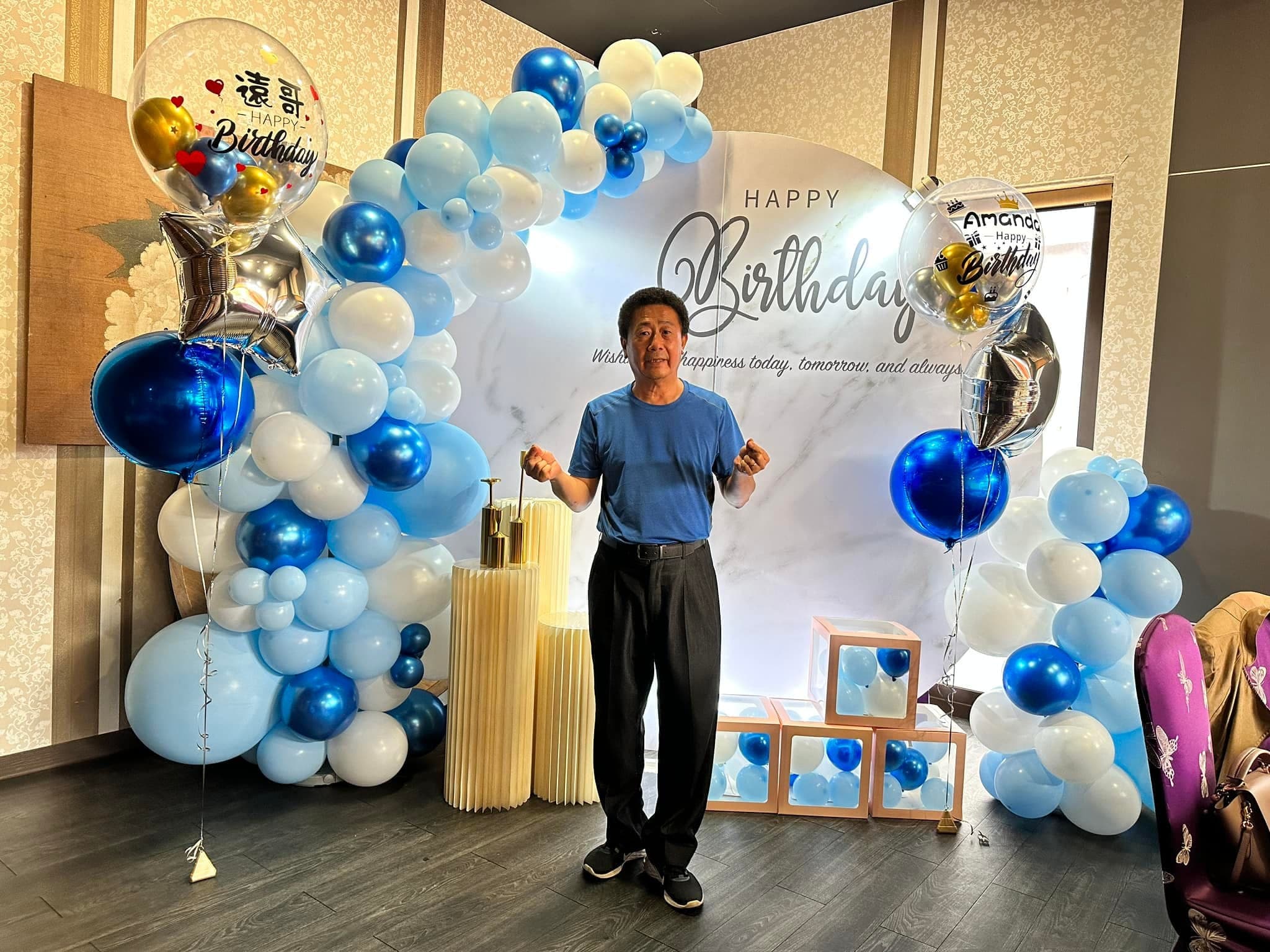 穿藍色上衣的男士站在藍白氣球拱門與生日背板前比手勢
