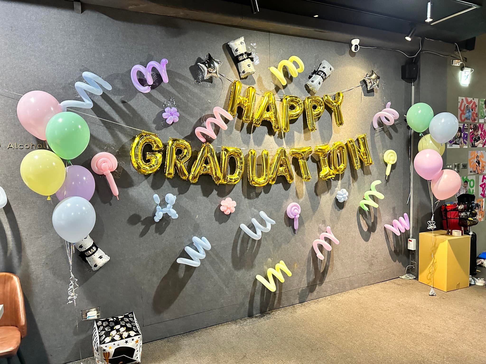 隱士國館畢業派對佈置，灰色布牆上貼著金色 HAPPY GRADUATION 字母與粉彩造型氣球