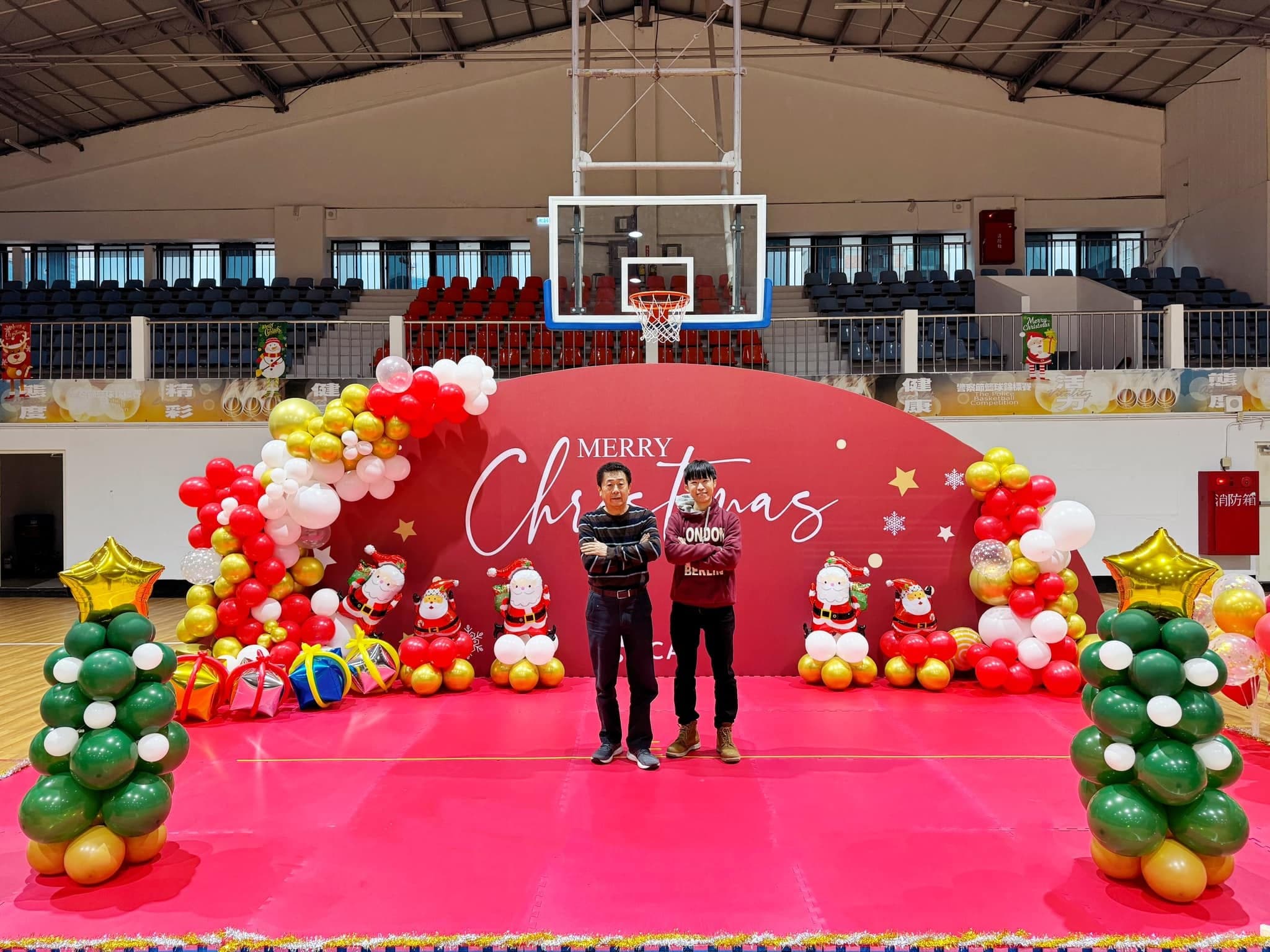 體育館舞台上的紅色 Merry Christmas 背板，兩側是紅白金氣球、聖誕老公公和綠金聖誕樹氣球柱，兩人站在中間