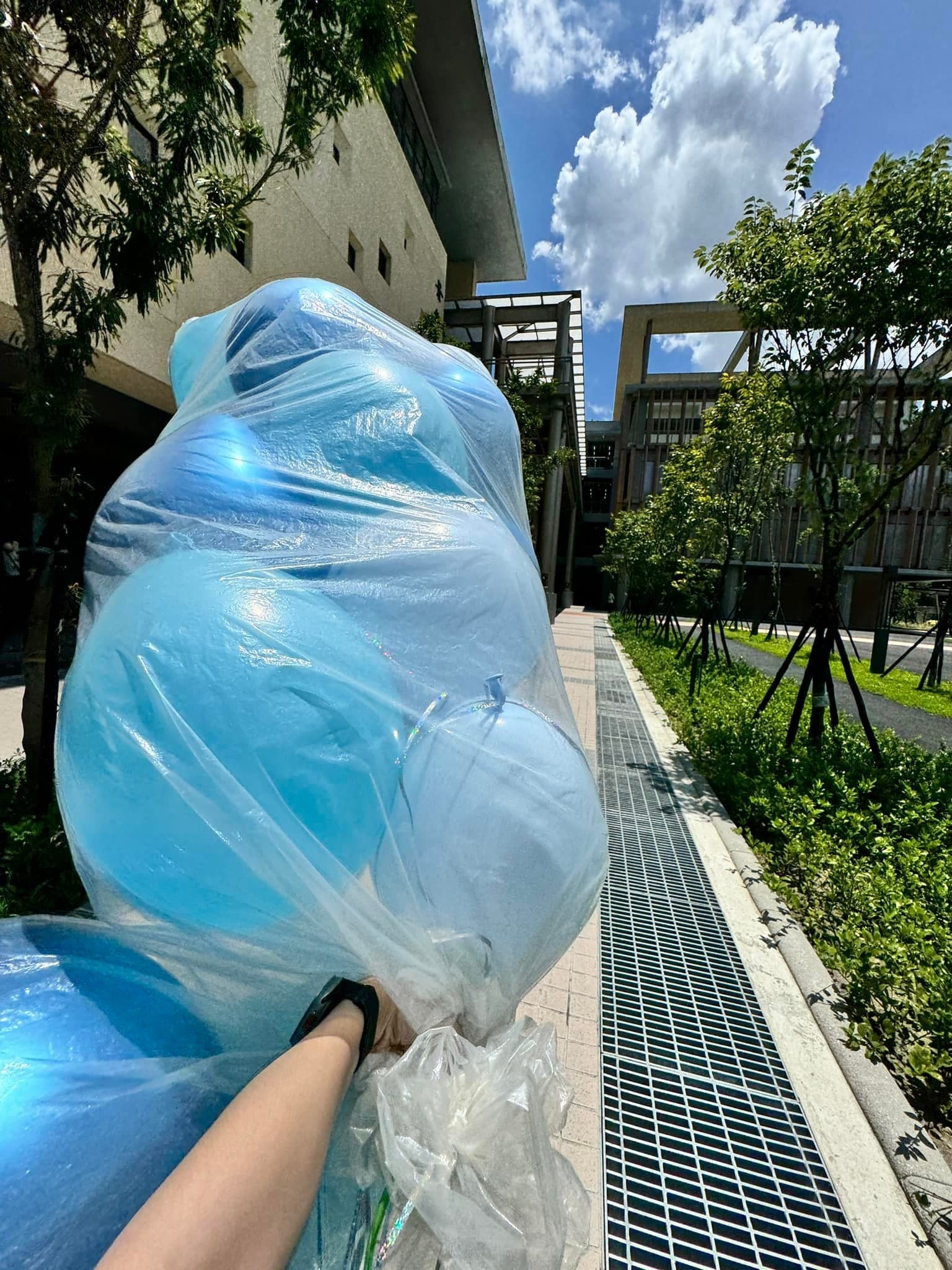 準備送往告別式的淺藍色氣球，用透明塑膠袋整袋包好，背景是藍天與步道