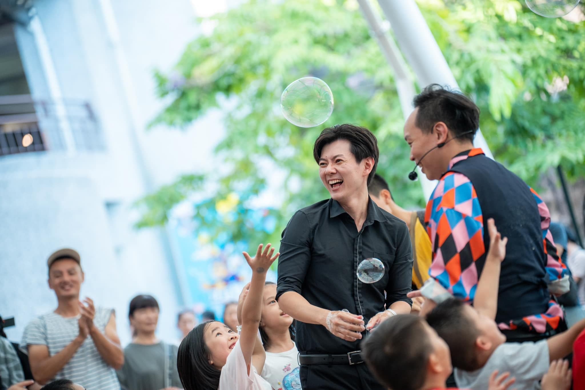 兩位表演者在台下吹出泡泡，孩子們伸手想抓住飄在空中的泡泡