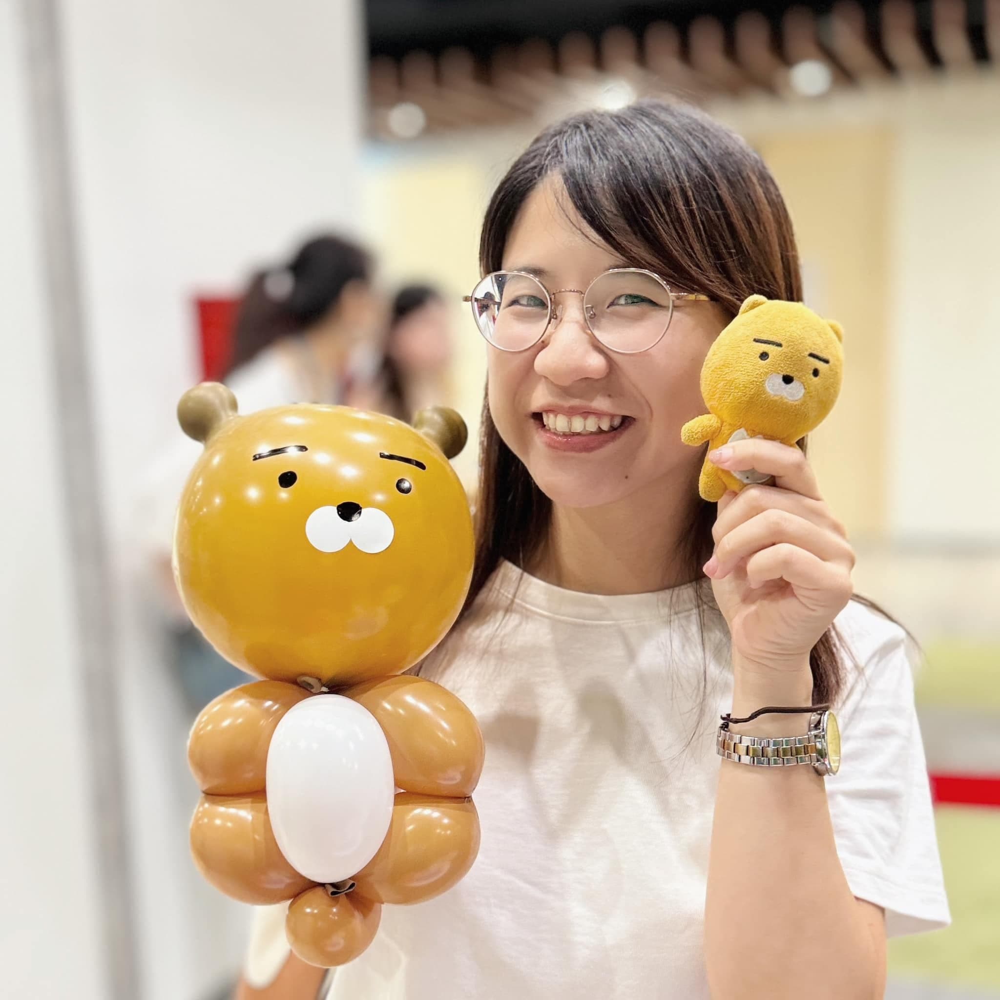 戴眼鏡的女生一手拿 LINE FRIENDS「Ryan」造型氣球，一手拿長得一樣的 Ryan 娃娃