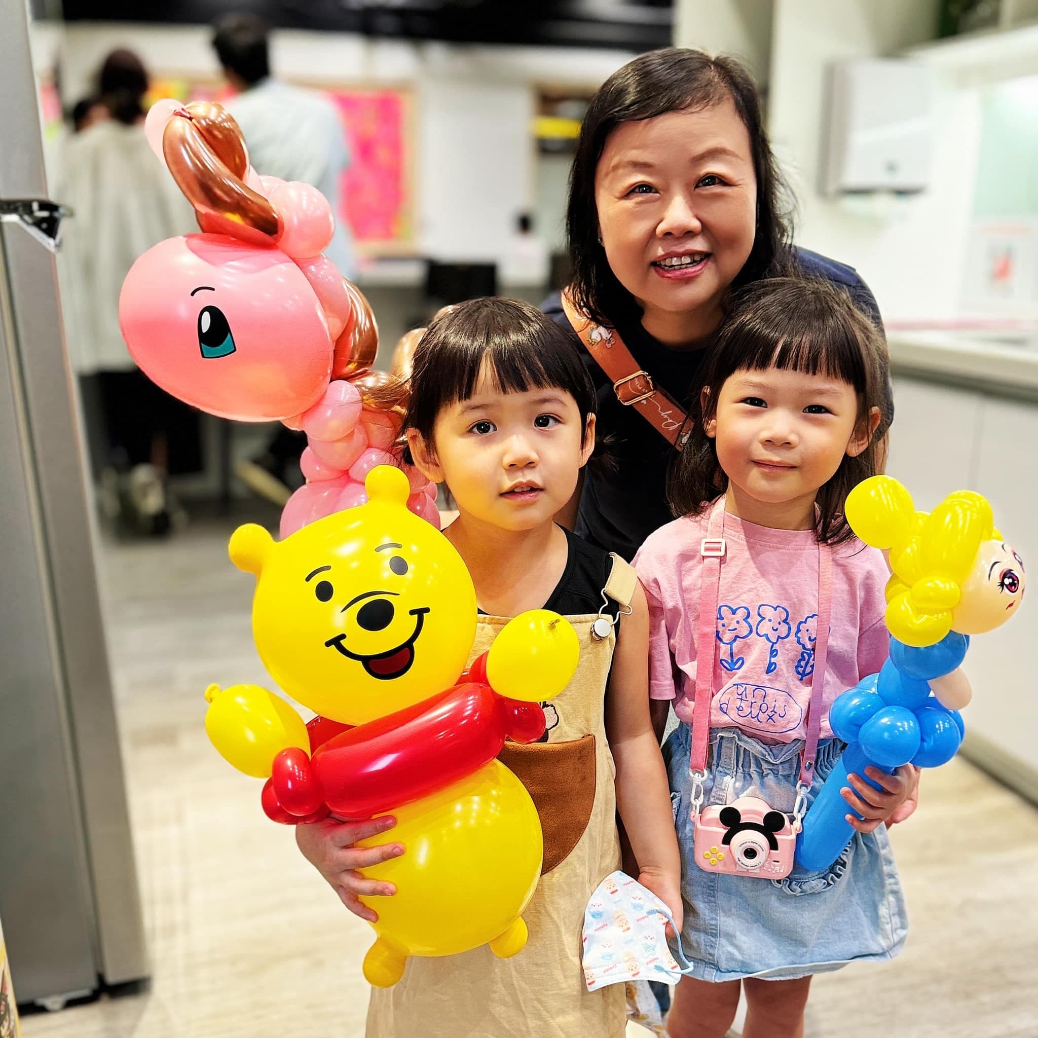 兩個小女孩和長輩合照，手上拿著小熊維尼、粉紅色小馬與藍色角色造型氣球