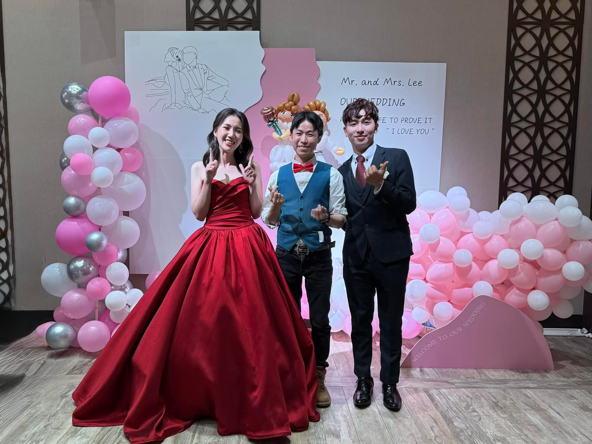 穿紅色禮服的新娘、西裝新郎和柯男站在粉紅白銀氣球拍照區前合照