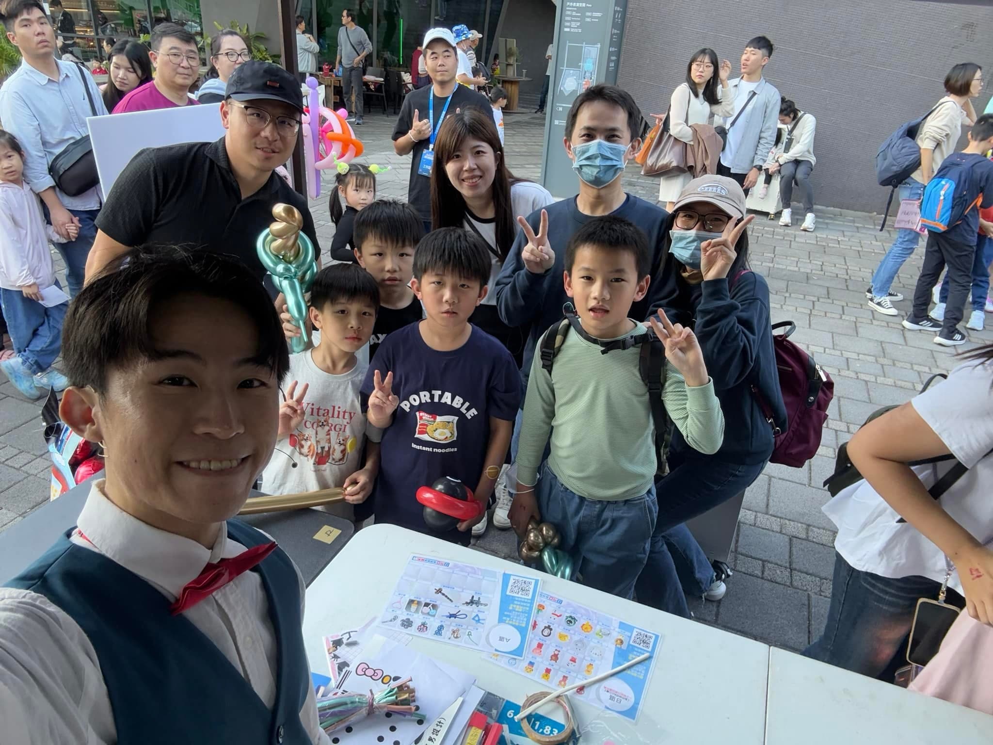 柯男在藝術節攤位前自拍，身後是拿著造型氣球比 YA 的小朋友與家長
