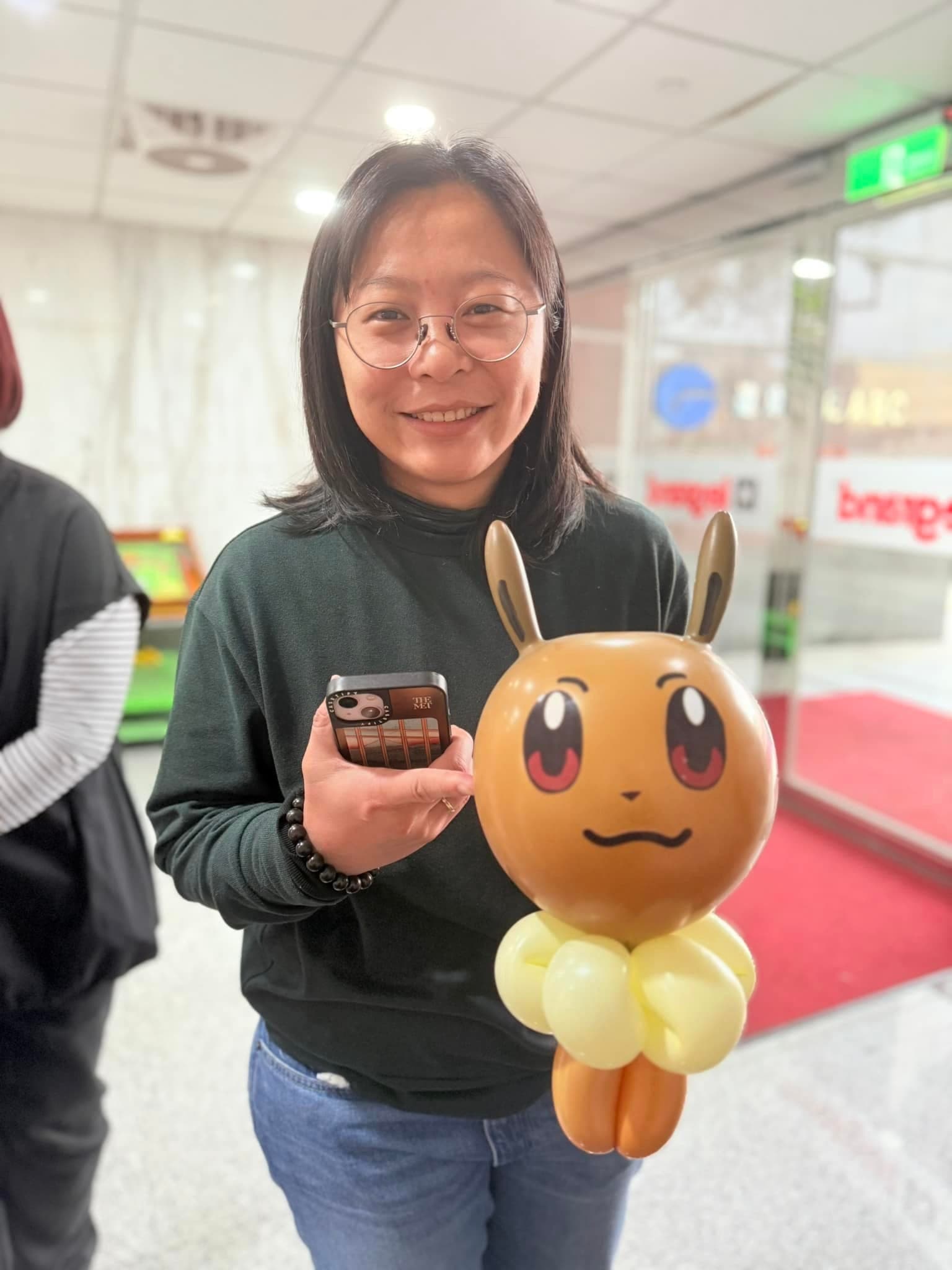 穿深綠色上衣的女士拿著咖啡色伊布造型氣球，脖子是奶油色圍毛